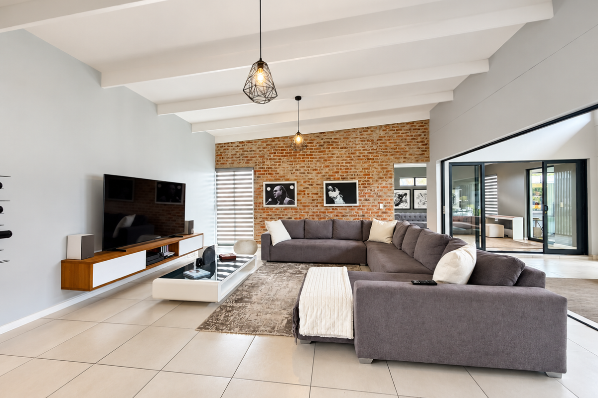 Spacious living room featuring an exposed brick wall, vaulted ceiling with exposed beams, large format floor tiles, and seamless access to the outdoor patio.