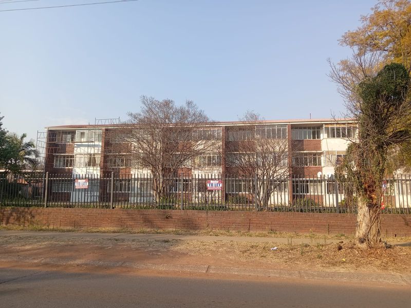 Front exterior view of a multi-story apartment building featuring brick facade, steel windows, and perimeter security fencing, currently undergoing exterior maintenance.
