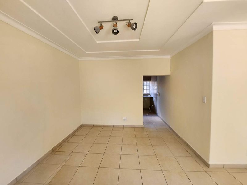 An empty, neutrally painted living room featuring tiled flooring, a decorative recessed ceiling, and modern track lighting, leading into an adjacent kitchen area.