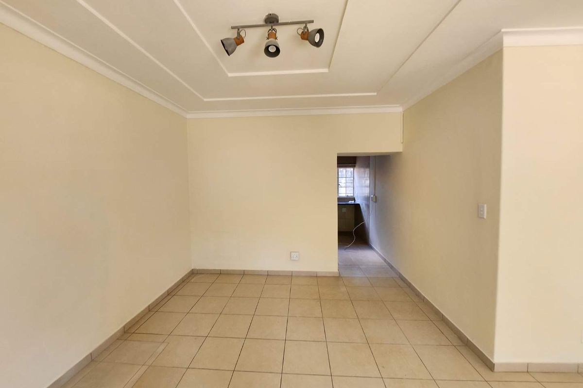 An empty, neutrally painted living room featuring tiled flooring, a decorative recessed ceiling, and modern track lighting, leading into an adjacent kitchen area.