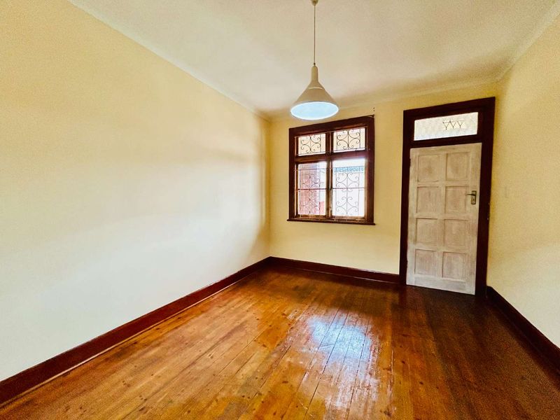 An empty bedroom featuring original hardwood flooring, timber window frames with security bars, and a classic timber door with a transom window.