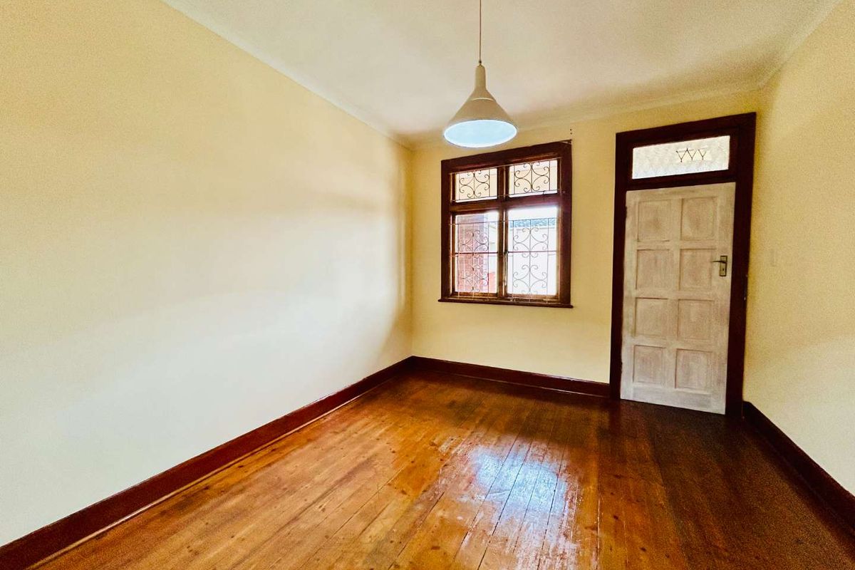 An empty bedroom featuring original hardwood flooring, timber window frames with security bars, and a classic timber door with a transom window.