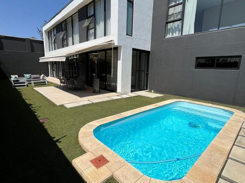 Modern rear exterior featuring a swimming pool, paved patio area, retractable awning, and artificial lawn set against a contemporary two-story home facade.