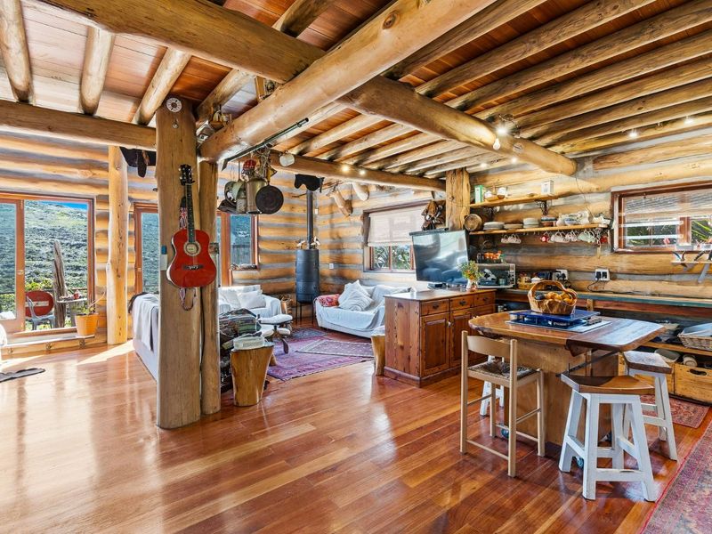 Rustic open-plan living area featuring exposed log walls, timber beam ceilings, hardwood flooring, and a wood-burning stove with access to outdoor space.