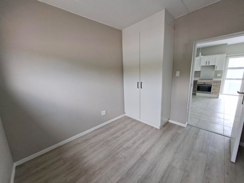 A clean, empty bedroom featuring modern laminate flooring and a built-in wardrobe, with a view through to the adjacent kitchen area.