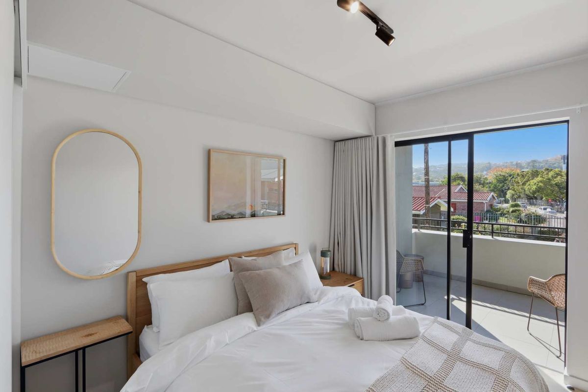 A bright bedroom featuring neutral tones, modern track lighting, and large sliding glass doors that open onto a private balcony with neighborhood views.