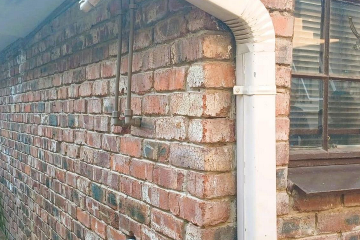 A narrow side passage featuring exposed brickwork, a gutter downpipe, and a window with a metal frame.