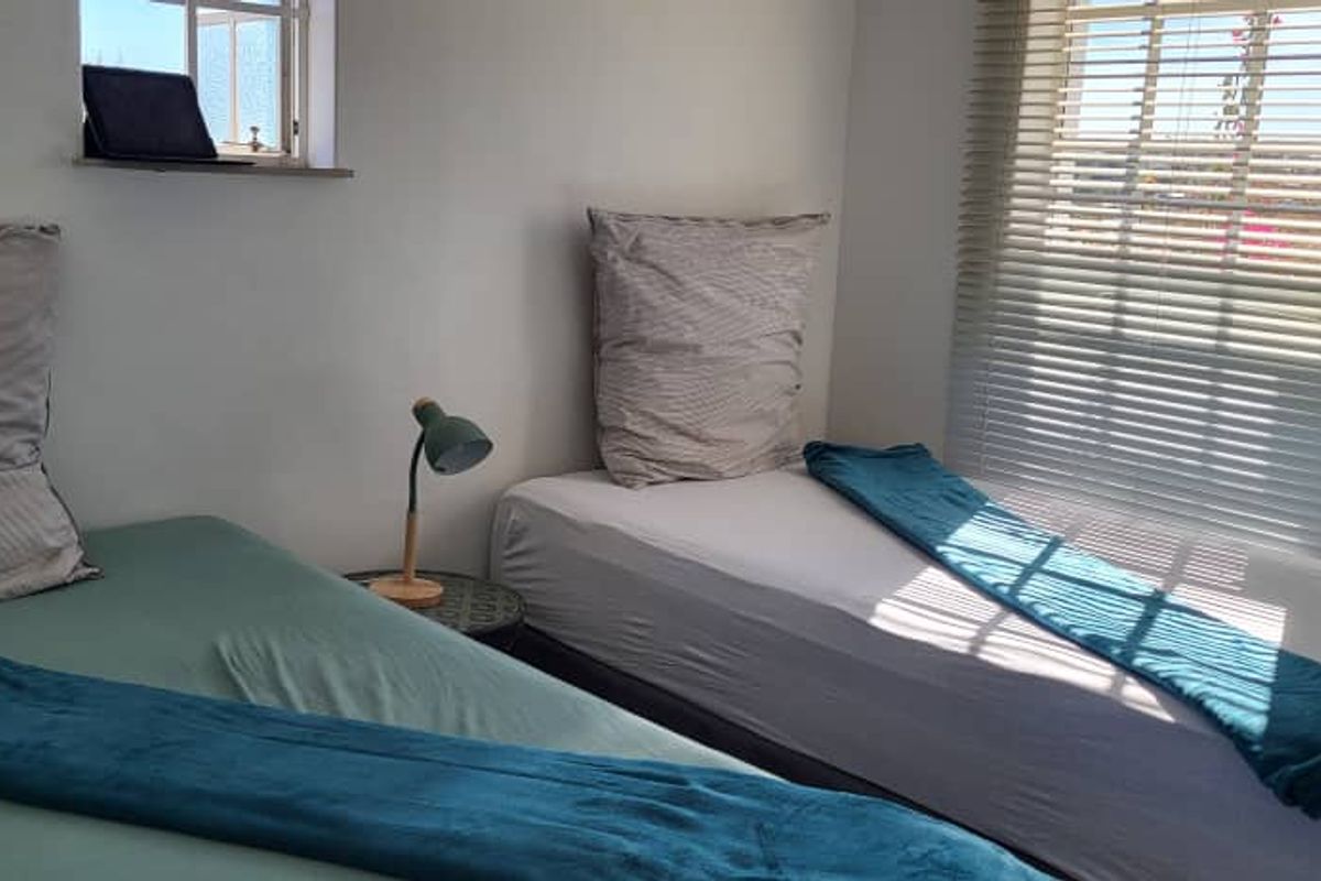 A compact bedroom featuring two single beds, white walls, and natural light provided by two windows fitted with venetian blinds.