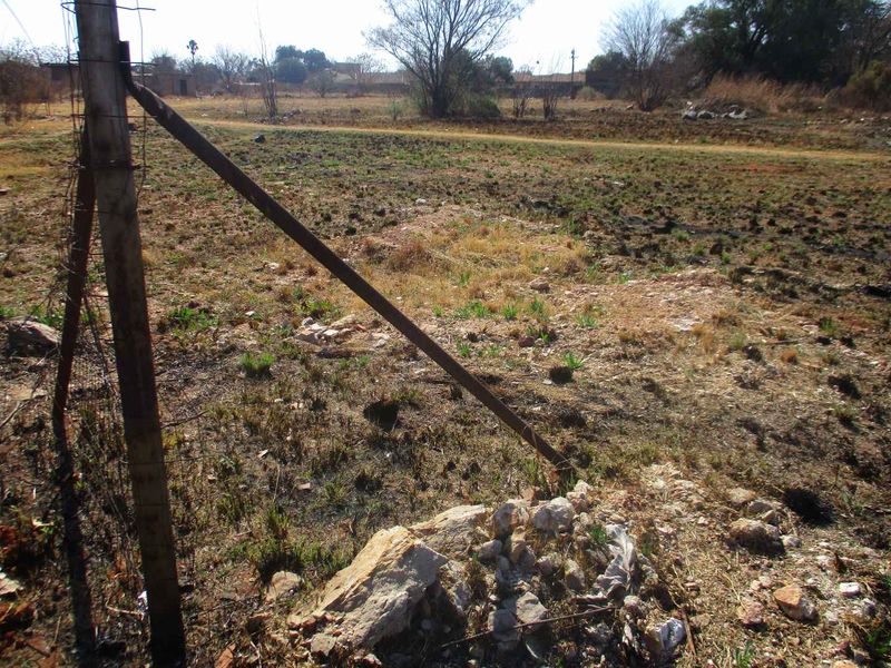 A view of a vacant residential plot featuring dry, unmaintained ground and damaged wire fencing.