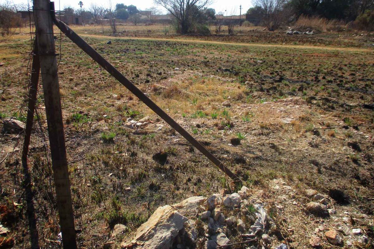 A view of a vacant residential plot featuring dry, unmaintained ground and damaged wire fencing.