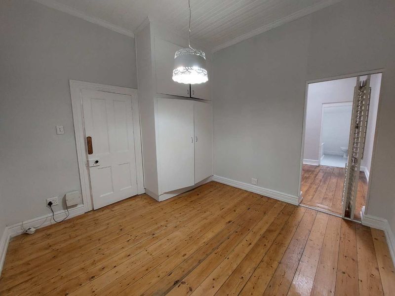 A bright, empty bedroom featuring polished timber flooring, built-in cupboards, and a classic beadboard ceiling, with an adjacent room visible through the doorway.