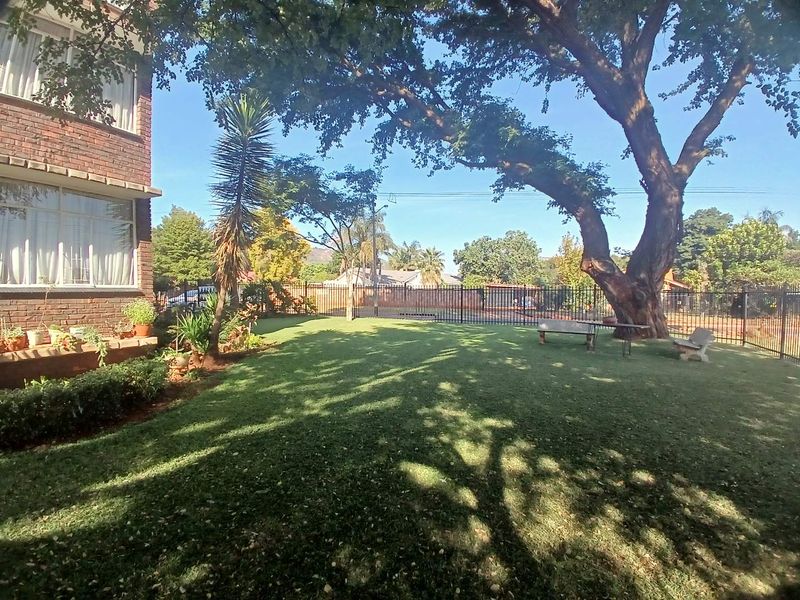 A well-maintained communal garden area featuring a large mature tree, manicured lawn, and secure metal perimeter fencing adjacent to the apartment building.