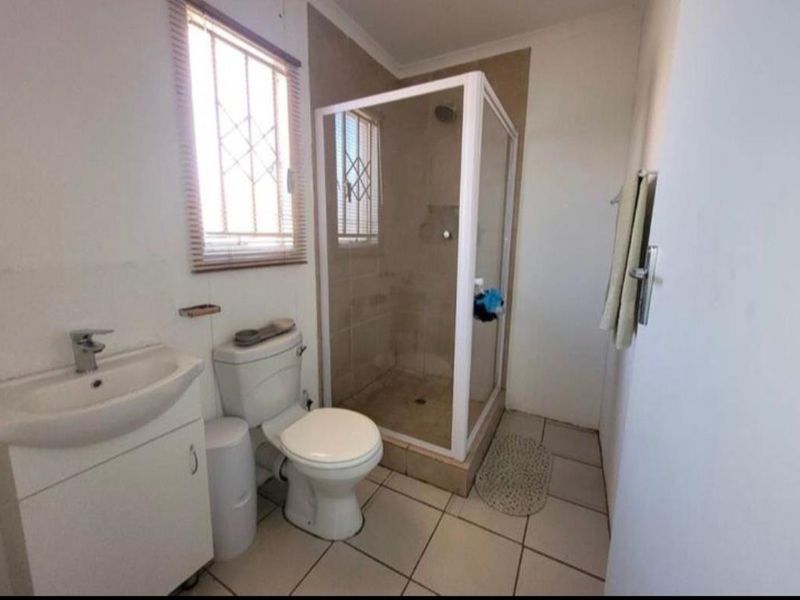A functional bathroom featuring a shower enclosure, vanity basin, and toilet with tiled flooring and natural light from a window with security bars.