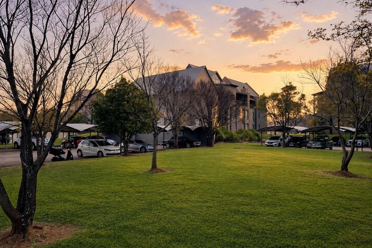 A view of the apartment complex grounds featuring a well-maintained lawn, mature trees, and covered parking facilities at sunset.