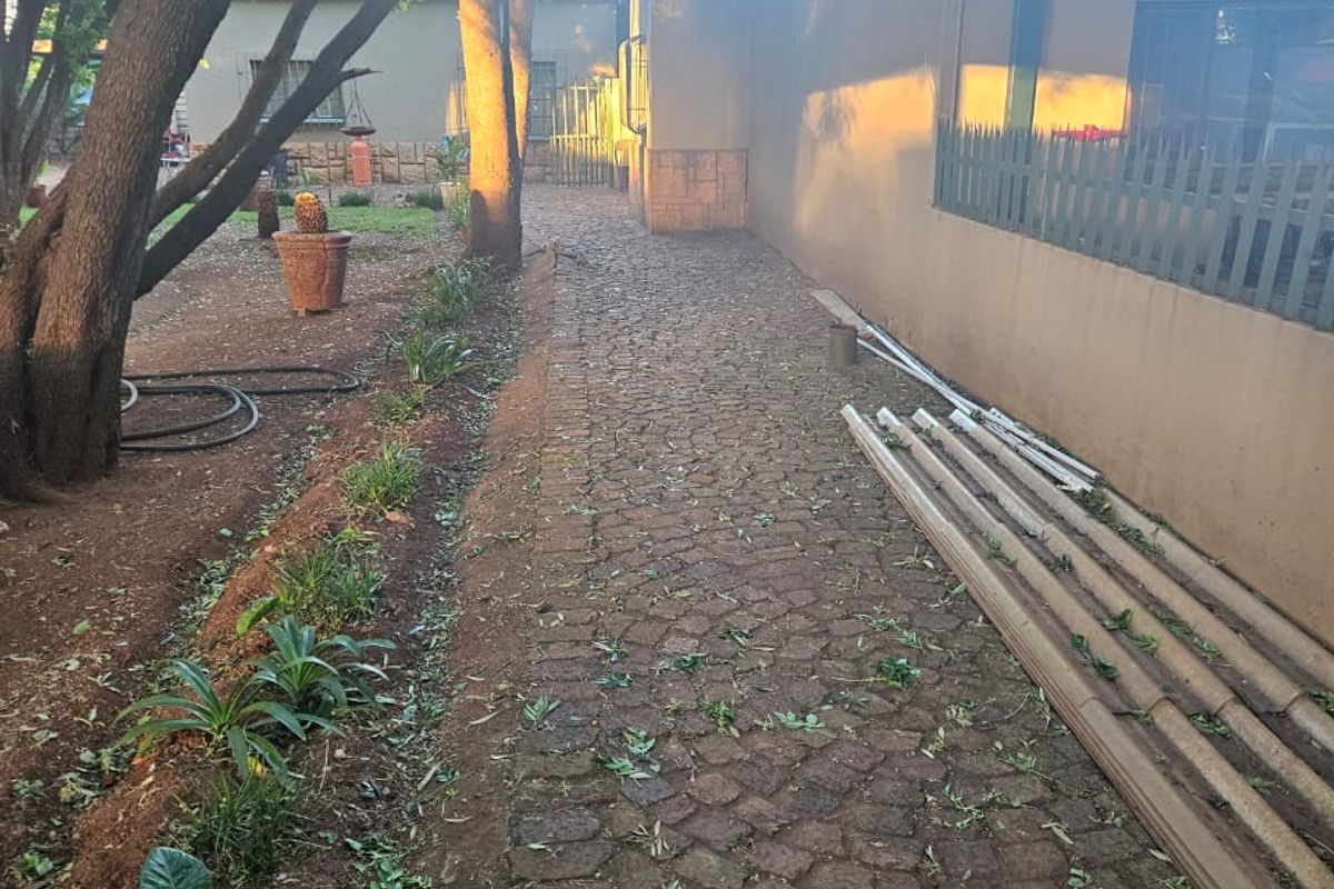 A rear exterior view of the cottage featuring a cobblestone pathway alongside the building, mature trees, and a metal security fence.