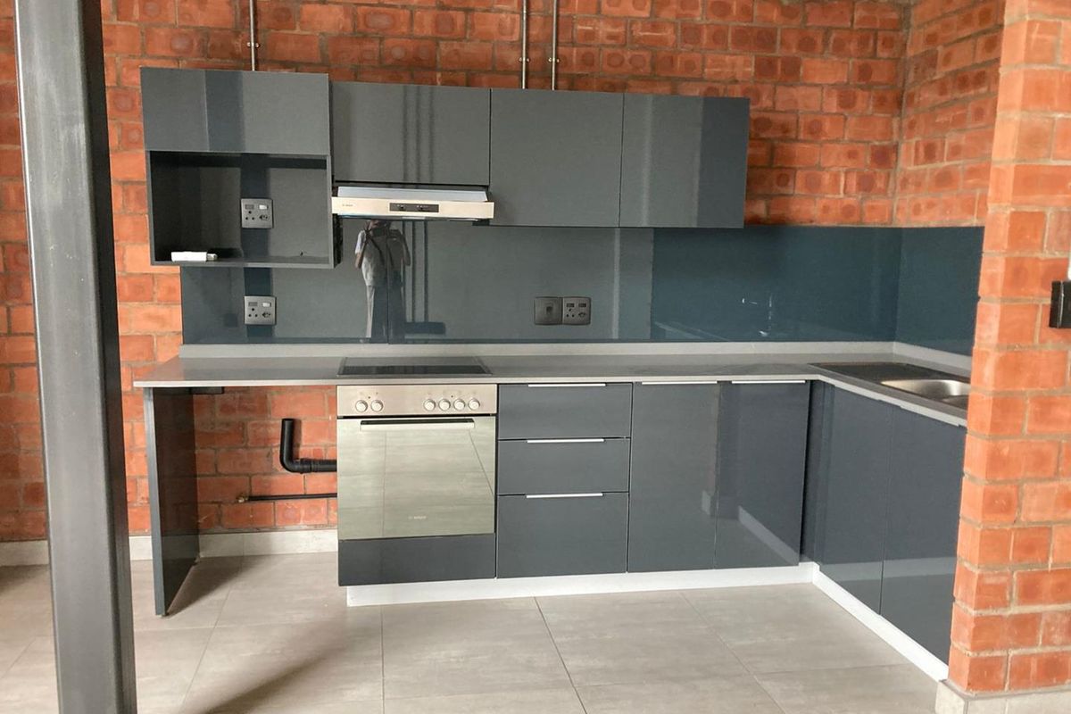 Modern kitchen featuring high-gloss grey cabinetry, integrated oven and hob, glass splashback, and industrial-style exposed brick walls.