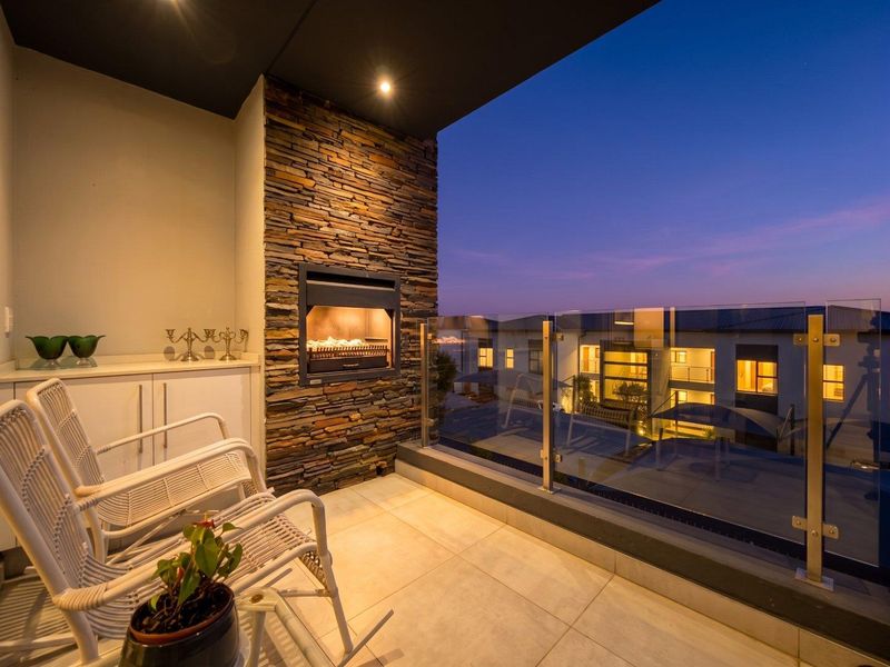 A modern covered balcony featuring a built-in stone-clad fireplace, glass balustrade, and tiled flooring, offering an inviting outdoor space with evening views.