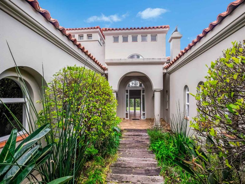 A Mediterranean-style home entrance featuring a terracotta tiled roof, stucco facade, arched architectural details, and a rustic timber sleeper walkway leading to the front door.