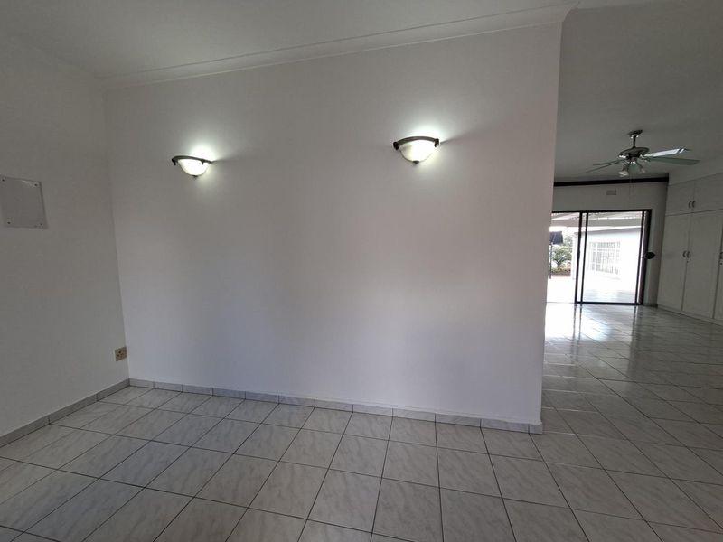 An empty living area featuring neutral ceramic floor tiles, wall-mounted lighting, and a ceiling fan, with a view through to an adjacent space.