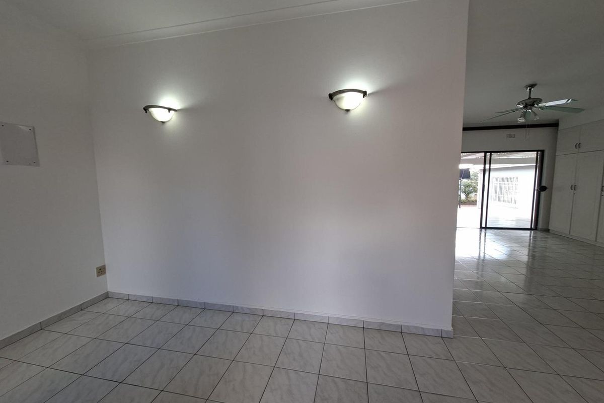 An empty living area featuring neutral ceramic floor tiles, wall-mounted lighting, and a ceiling fan, with a view through to an adjacent space.