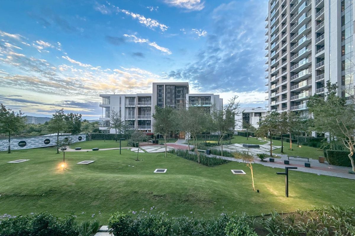 A view of the landscaped communal garden area and modern apartment building complex, featuring manicured lawns, paved pathways, and integrated outdoor lighting.