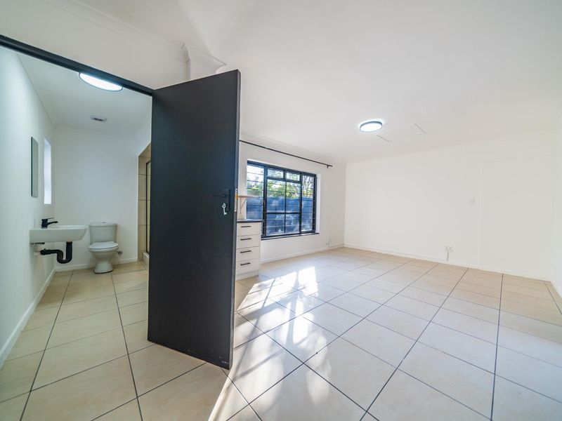 A bright, empty bedroom featuring tiled flooring, a large steel-framed window, and an en-suite bathroom with a vanity, toilet, and shower.