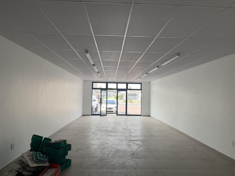 An empty open-plan retail space featuring a suspended ceiling, fluorescent lighting, tiled flooring, and a large glass storefront entrance.