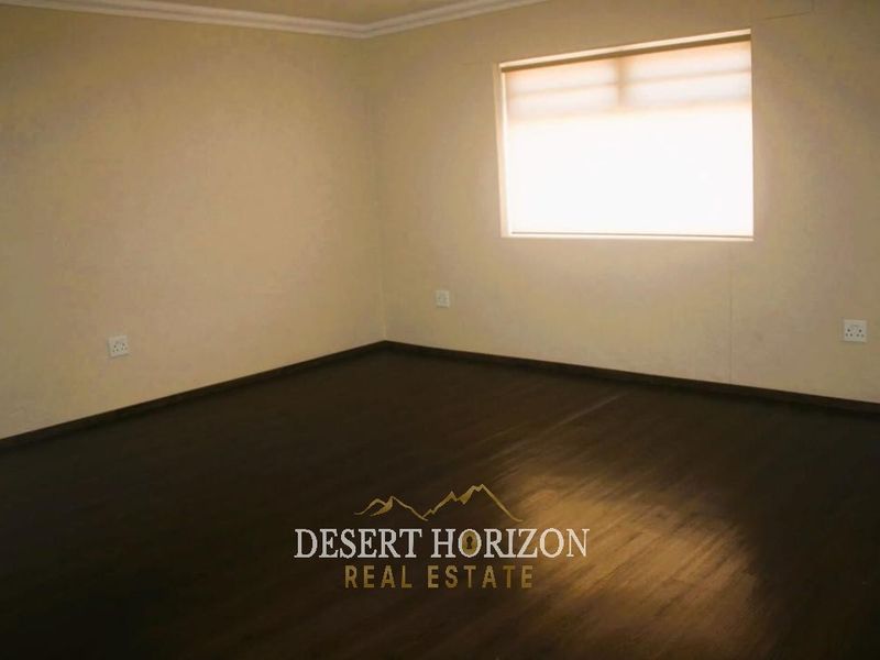 A spacious, empty living area featuring dark laminate flooring, neutral walls, and a window fitted with a roller blind.