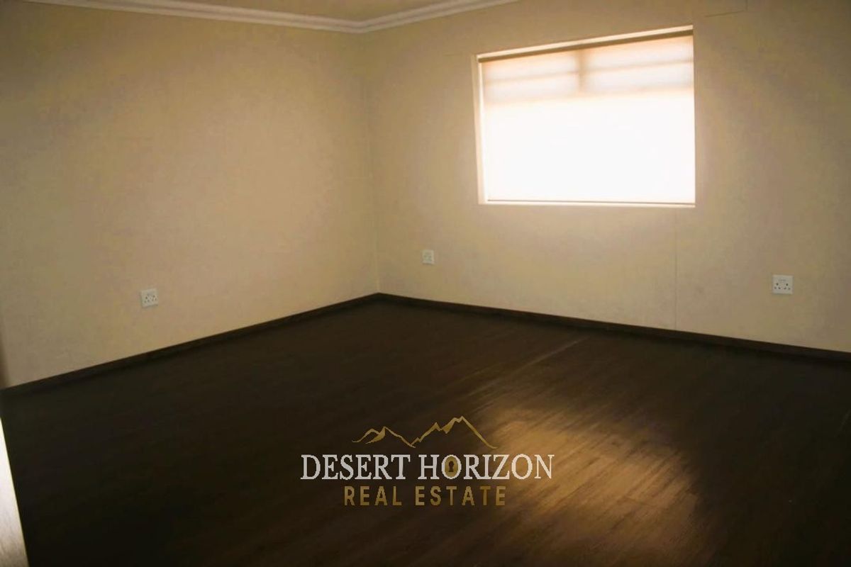 A spacious, empty living area featuring dark laminate flooring, neutral walls, and a window fitted with a roller blind.