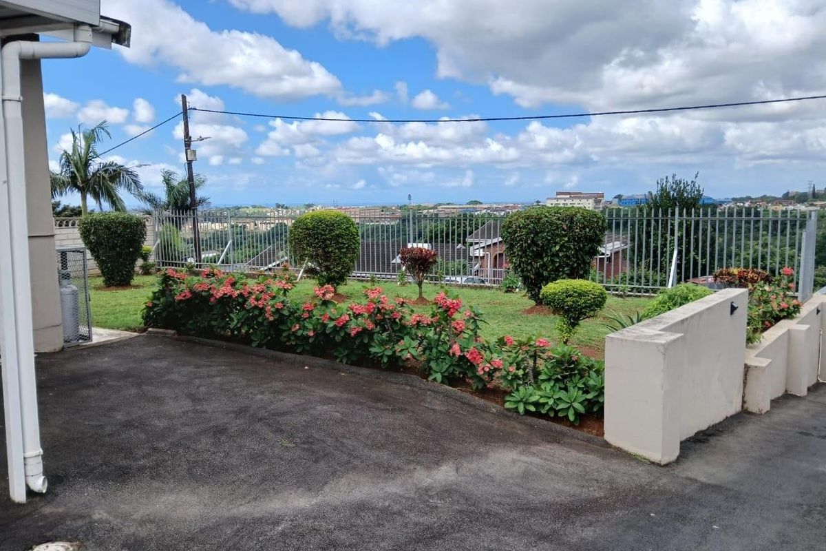 A well-maintained rear exterior area featuring an asphalt driveway, landscaped garden beds, and metal security fencing with an elevated neighborhood view.