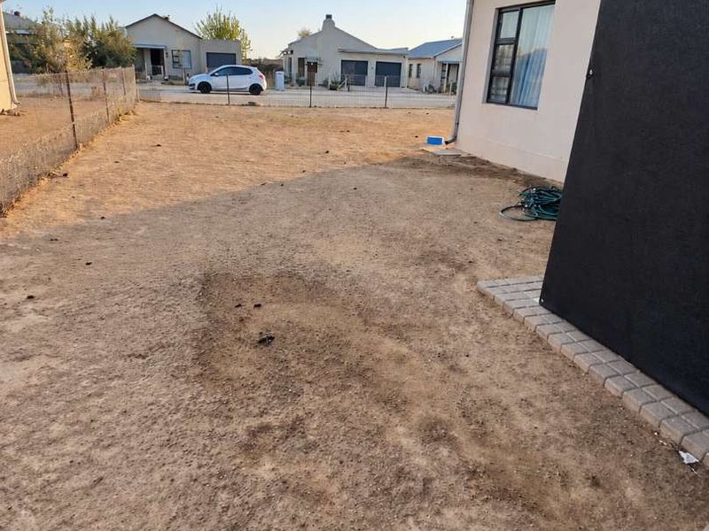 A rear yard area featuring a large open space with sparse ground cover, perimeter fencing, and a paved border along the building exterior.