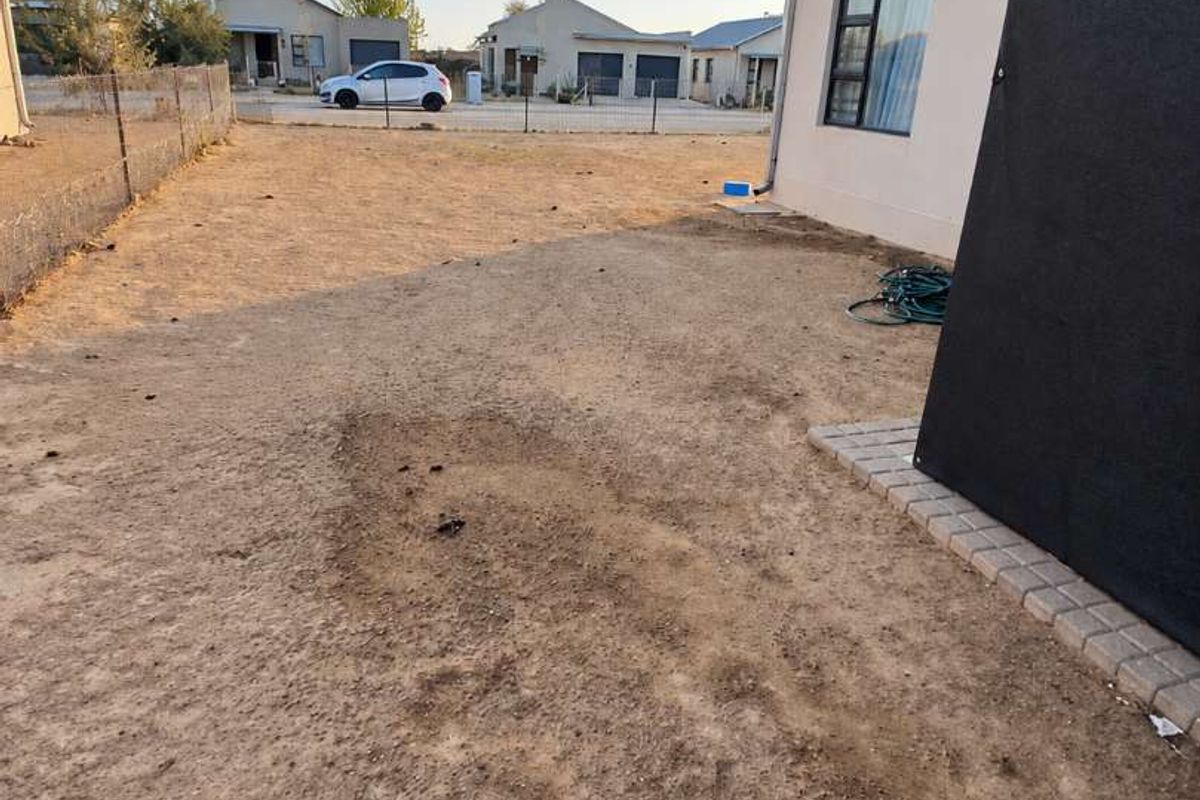 A rear yard area featuring a large open space with sparse ground cover, perimeter fencing, and a paved border along the building exterior.