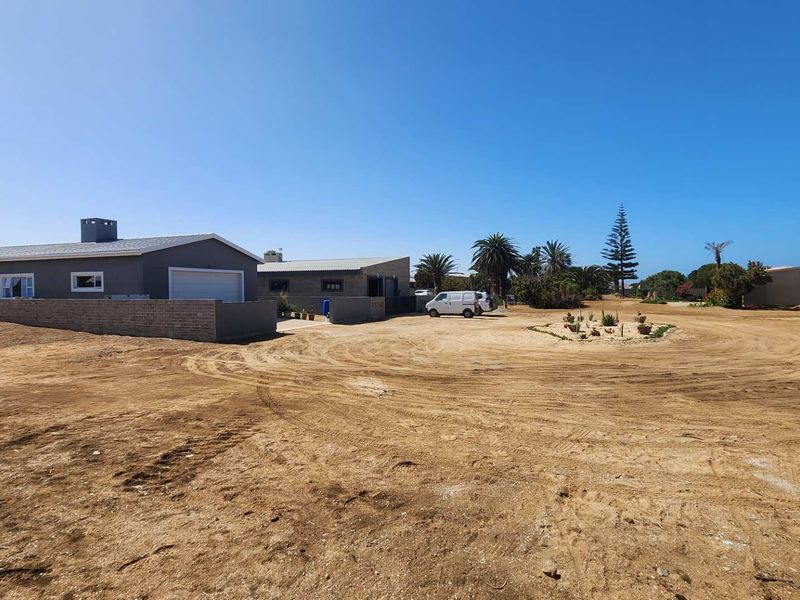 A wide-angle view of a residential property featuring a large, unpaved sandy yard, a brick boundary wall, and a garage structure.