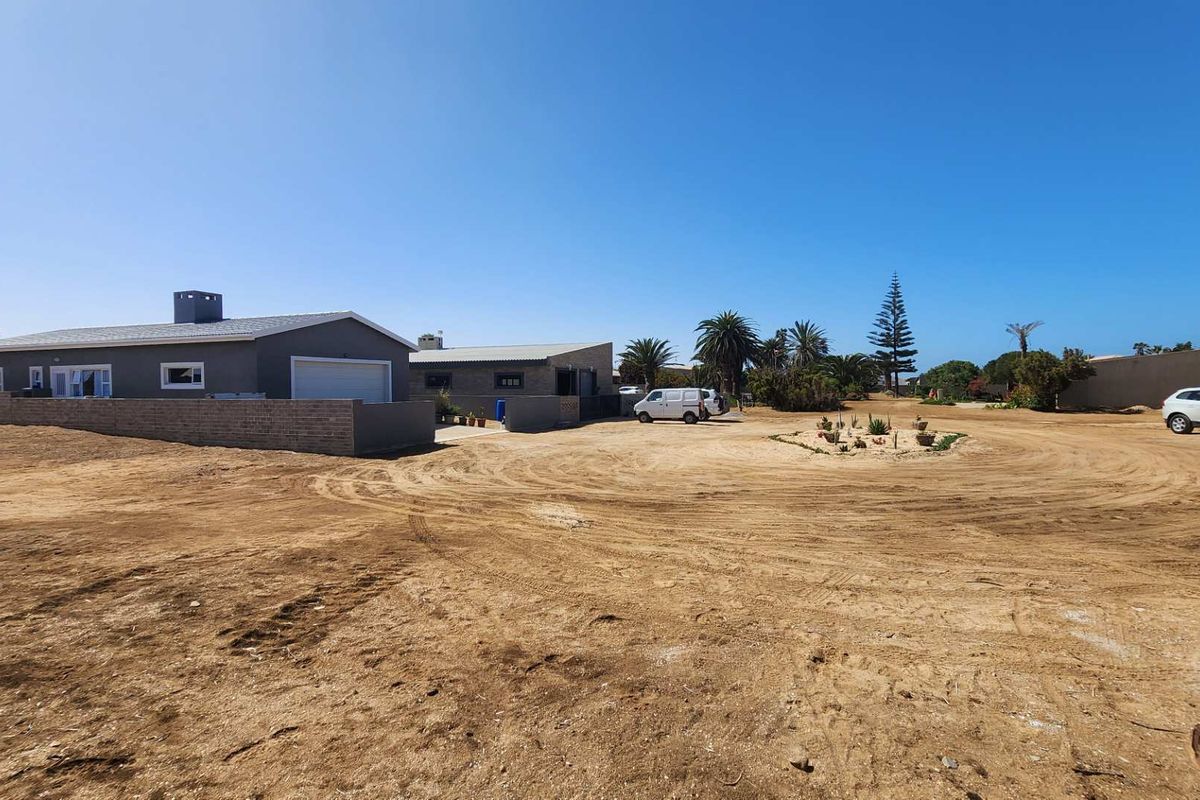 A wide-angle view of a residential property featuring a large, unpaved sandy yard, a brick boundary wall, and a garage structure.