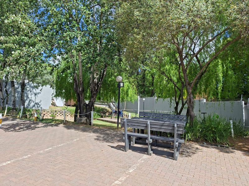 A paved outdoor area featuring mature trees, perimeter walling with electric fencing, and garden lighting in a communal or residential setting.