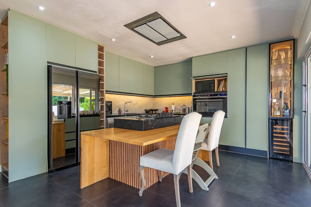 Modern kitchen featuring sage green cabinetry, a central island with a wooden breakfast bar, integrated appliances, and sleek dark stone flooring.