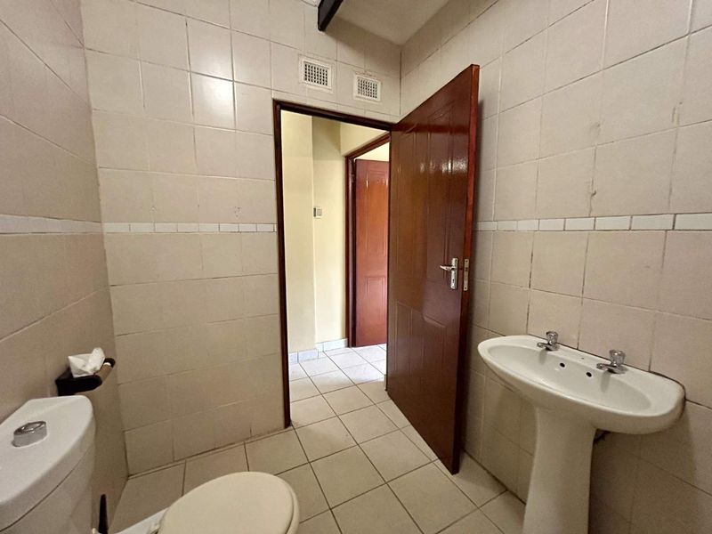 A functional bathroom featuring a pedestal washbasin, toilet, and full-height ceramic wall tiling, requiring cosmetic updates to improve its dated appearance.