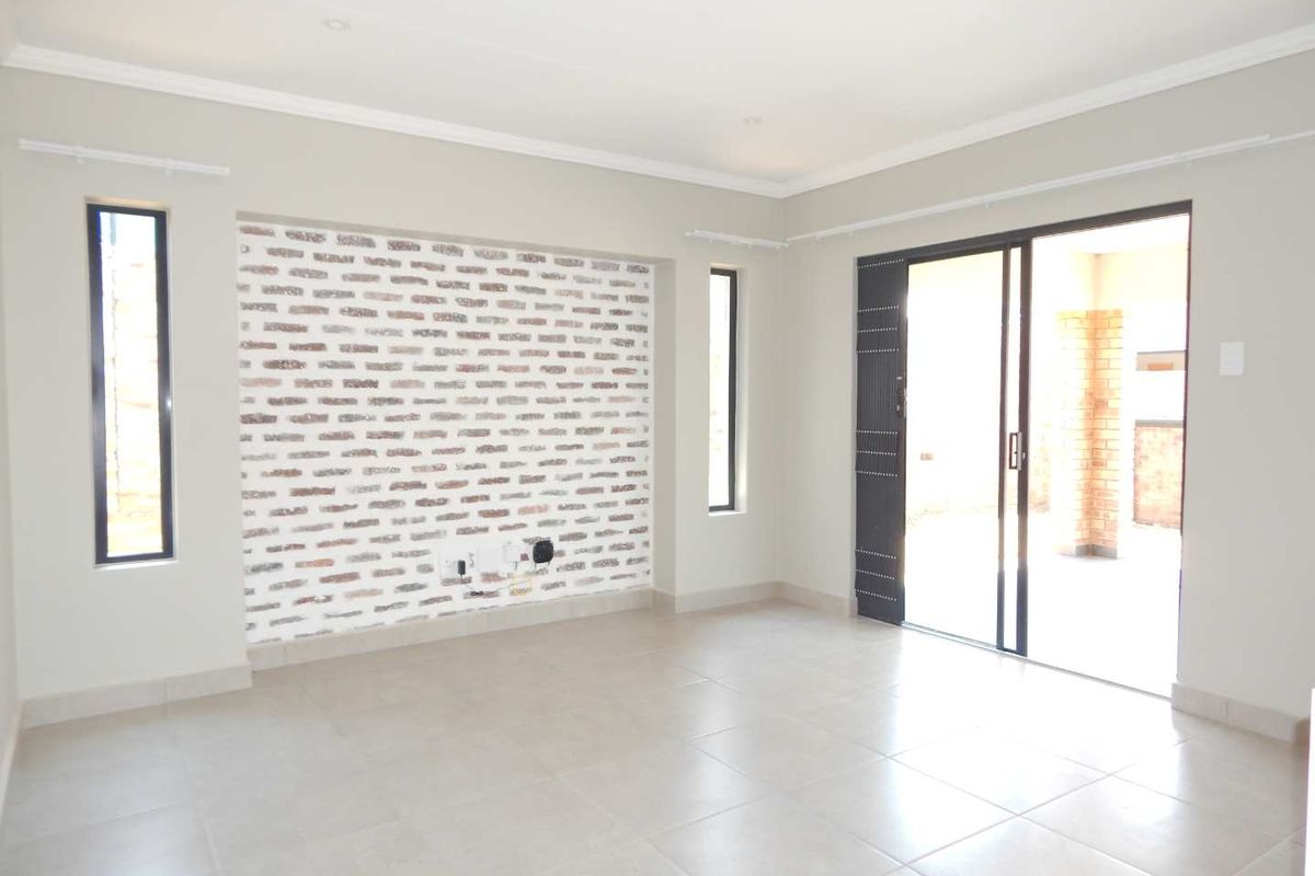 A bright, empty living room featuring a decorative exposed brick wall, neutral tiled flooring, and sliding glass doors leading to an outdoor patio area.
