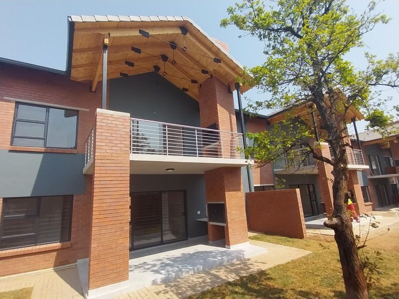 Rear view of a modern townhouse featuring a brick facade, covered patio with built-in braai, and an upper-level balcony with stainless steel railings.