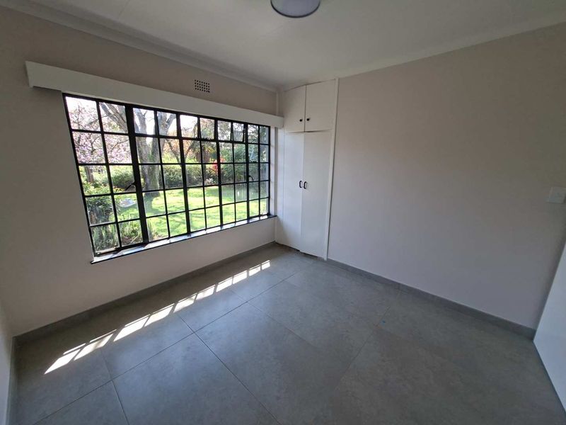 A bright, empty bedroom featuring large steel-framed windows overlooking the garden, neutral tiled flooring, and built-in storage cupboards.
