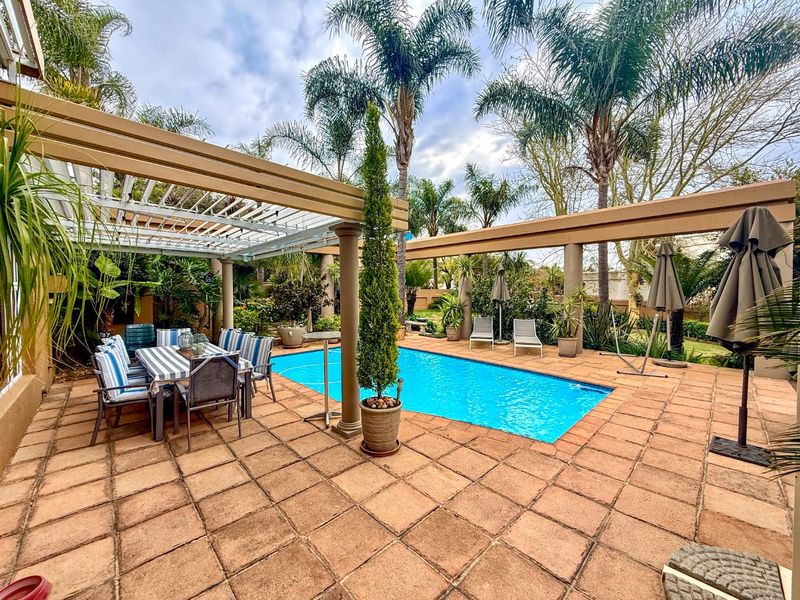 Outdoor pool area featuring a tiled patio, a large pergola structure with concrete columns, and established garden surroundings.