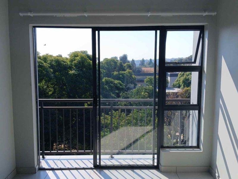 A bright, empty room featuring a sliding glass door that opens onto a balcony with a metal balustrade and scenic tree-lined views.