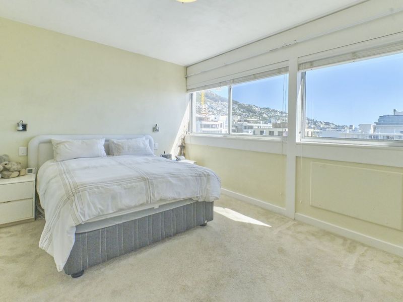 A bright bedroom featuring large windows with fitted blinds, carpeted flooring, and wall-mounted lighting, offering a pleasant view of the surrounding area.