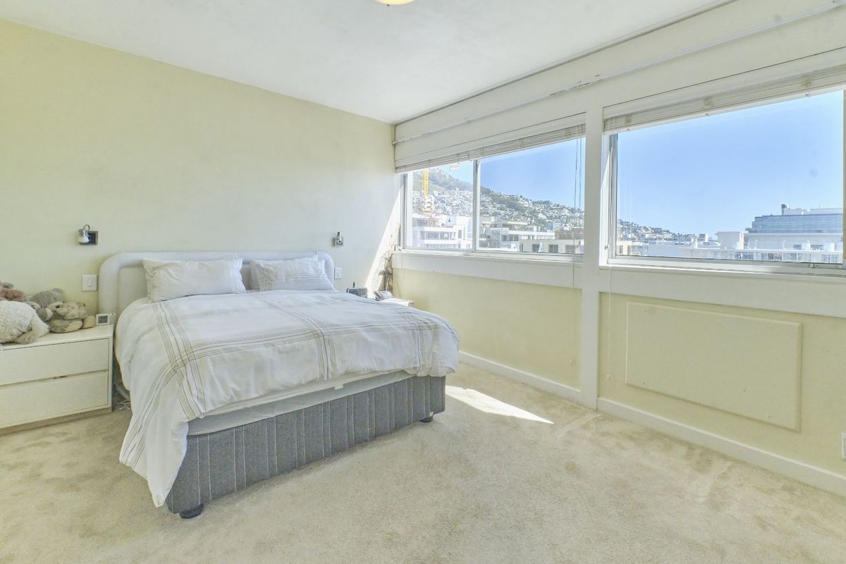 A bright bedroom featuring large windows with fitted blinds, carpeted flooring, and wall-mounted lighting, offering a pleasant view of the surrounding area.