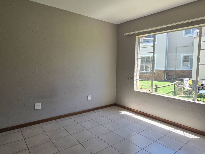 An empty bedroom featuring tiled flooring, neutral wall paint, and large windows fitted with security bars, offering a view of the complex grounds.