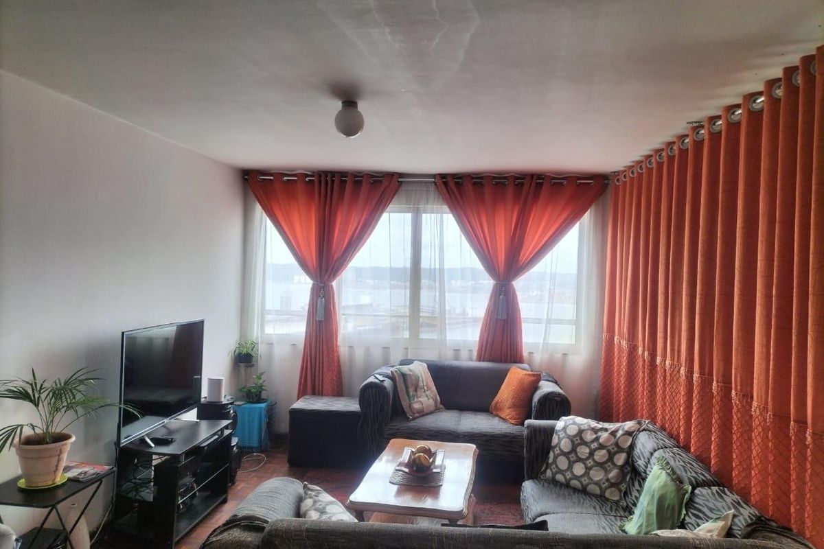 A living room featuring parquet flooring and a large window with natural light. Note visible water staining on the ceiling.