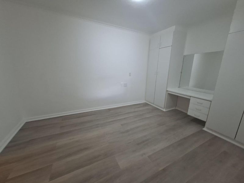 A bright, empty bedroom featuring modern laminate flooring, built-in cupboards, and a integrated vanity unit with a mirror.