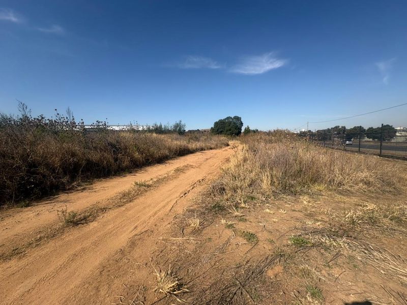 An undeveloped industrial land parcel featuring a dirt access track, perimeter fencing, and natural vegetation under a clear sky.