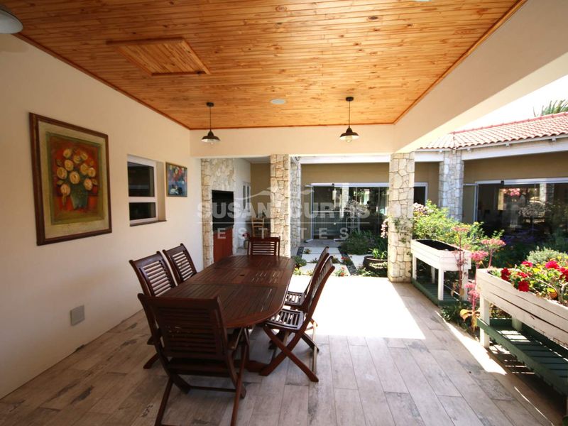 A spacious covered patio featuring a timber ceiling, stone-clad pillars, and a built-in braai, overlooking a landscaped garden area.