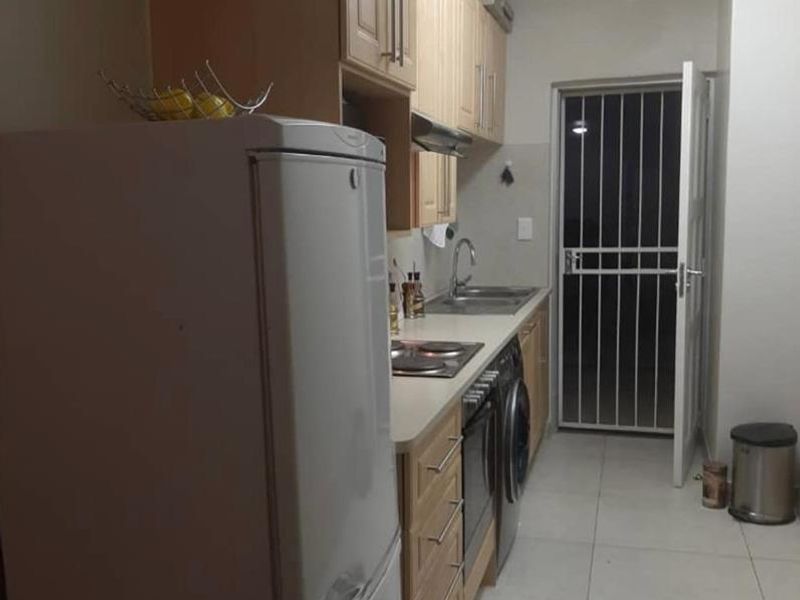 A functional galley-style kitchen featuring light wood cabinetry, built-in oven and hob, stainless steel sink, and tiled flooring with direct access to an exterior door.